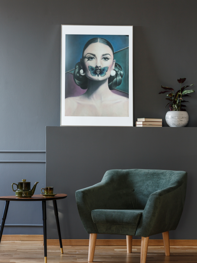 Pastel drawing of a woman with a butterfly over her mouth in a black frame stading on a dark grey shelf net to some books and a vase in a room with a dark green armchair and a coffee table