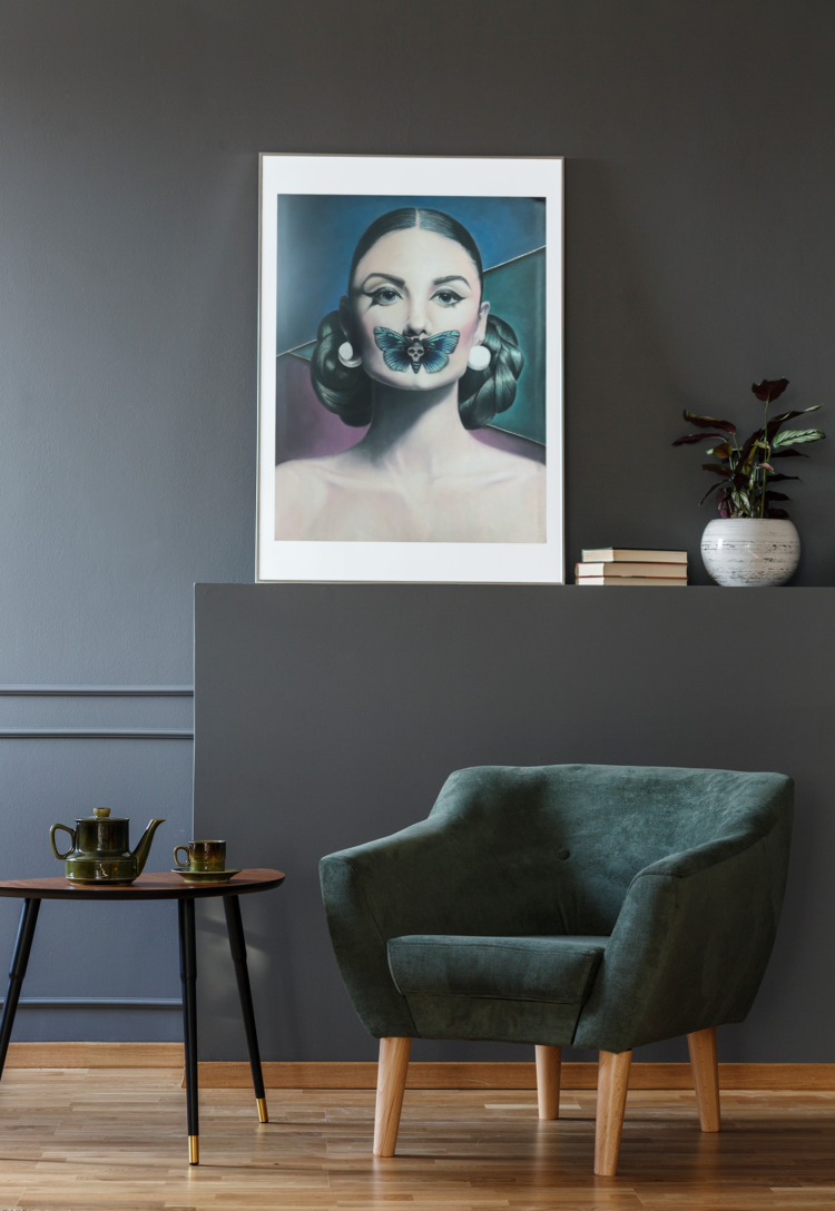 Pastel drawing of a woman with a butterfly over her mouth in a black frame stading on a dark grey shelf net to some books and a vase in a room with a dark green armchair and a coffee table
