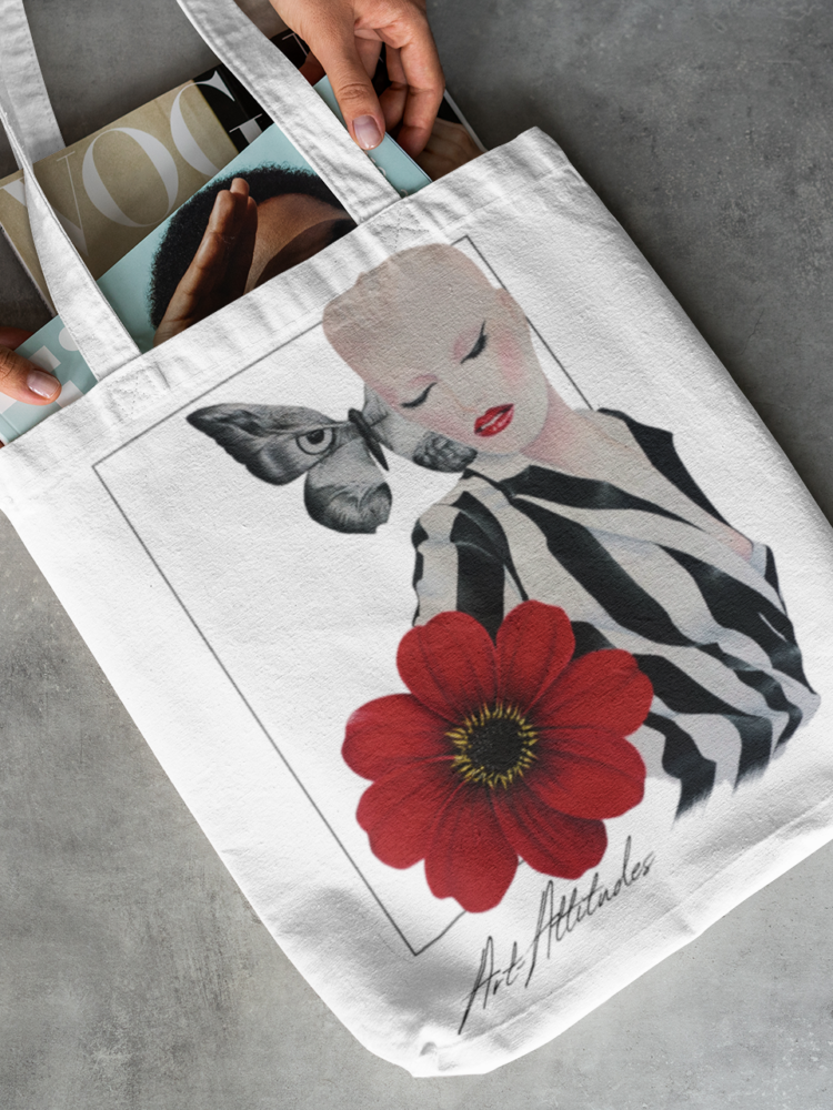 White tote bag laying on the floor with a motive of a woman in a balck and white striped shirt and a big red flower on her right side and a big butterfly next to her head