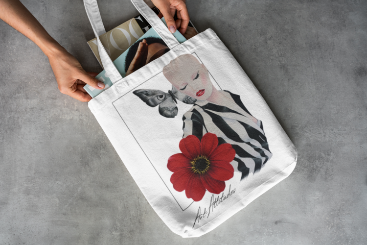 White tote bag laying on the floor with a motive of a woman in a balck and white striped shirt and a big red flower on her right side and a big butterfly next to her head