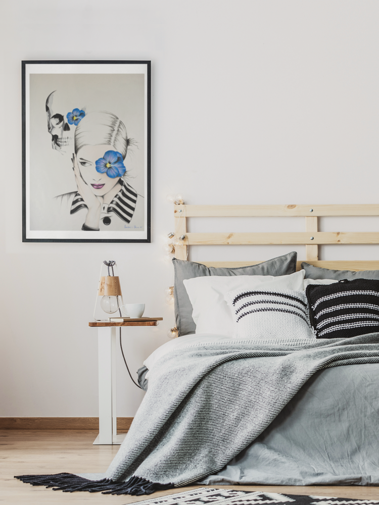 Pastel drawing of woman with blue flower in frent of her left eye in a white shirt with a striped collar and a skull in the bagground with a blue flowe on it's left eye in a black frame hanging on the wall next to a bed